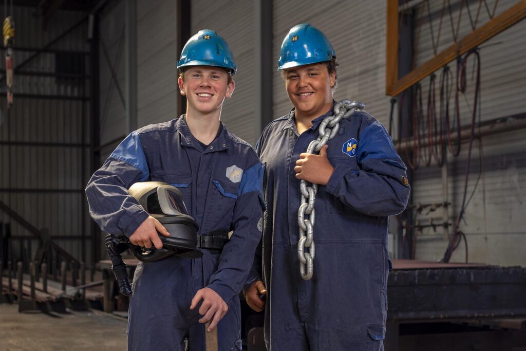 Twee schoolverlaters in overall en met helm van Nieuwenhuijsen Scheepsbouw staan in hun loods