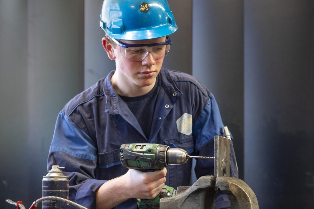 Schoolverlater in kleding van Nieuwenhuijsen Scheepsbouw heeft een boor in zijn hand en boort in een stuk staal.